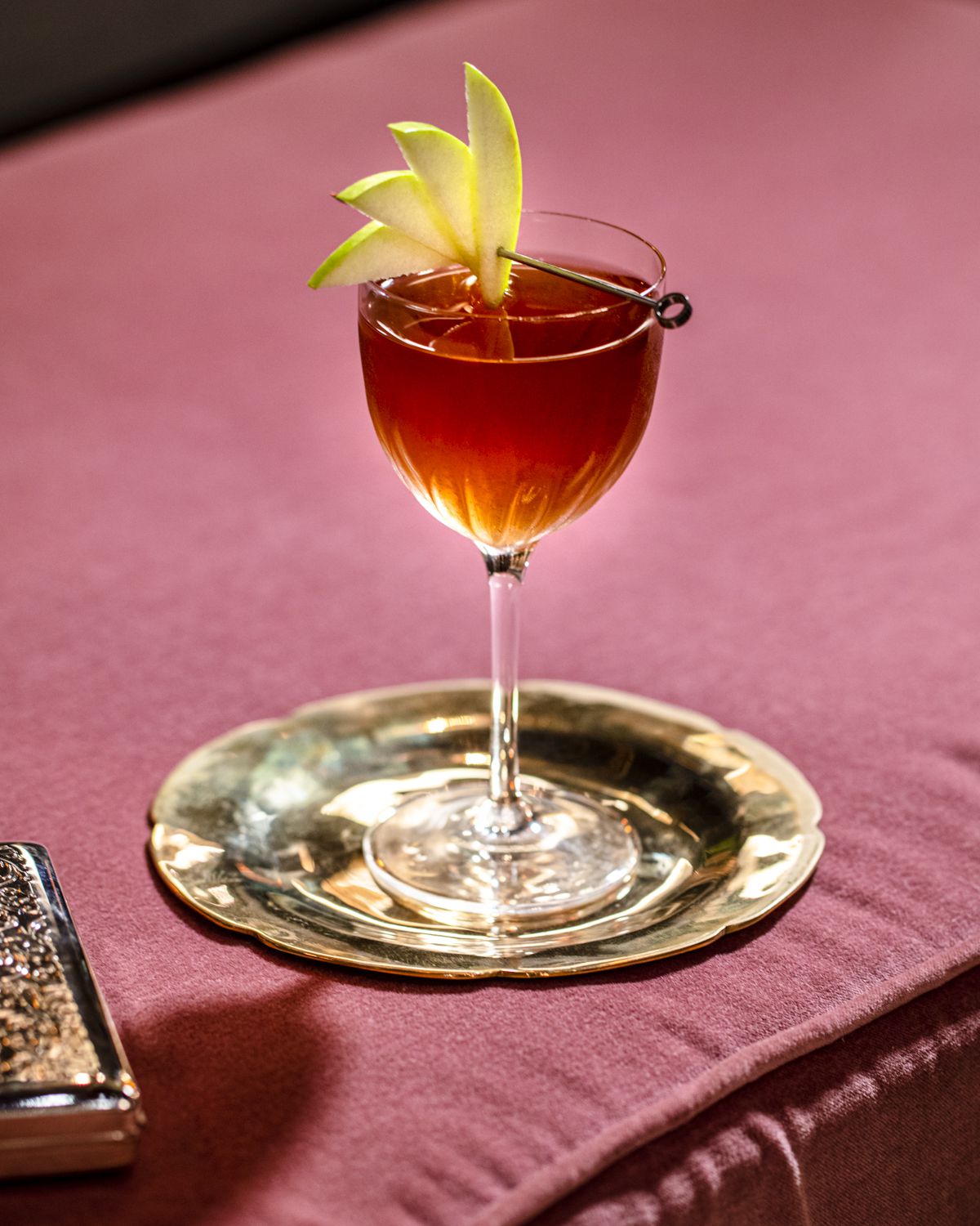A small dark cocktail in a glass with sliced apple garnish.