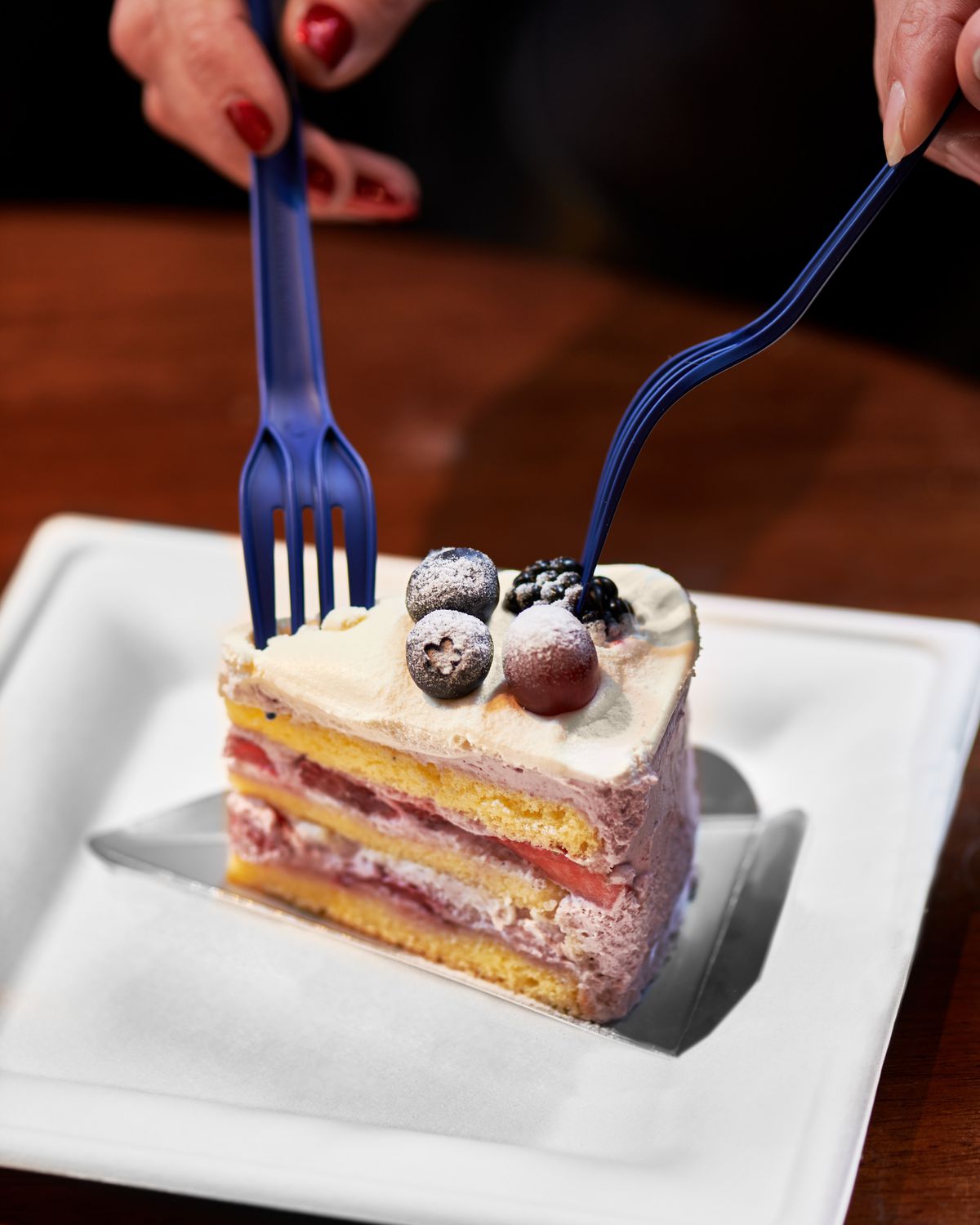 Two plastic forks stab into a slice of cake with berries on top and layers of berry filling.