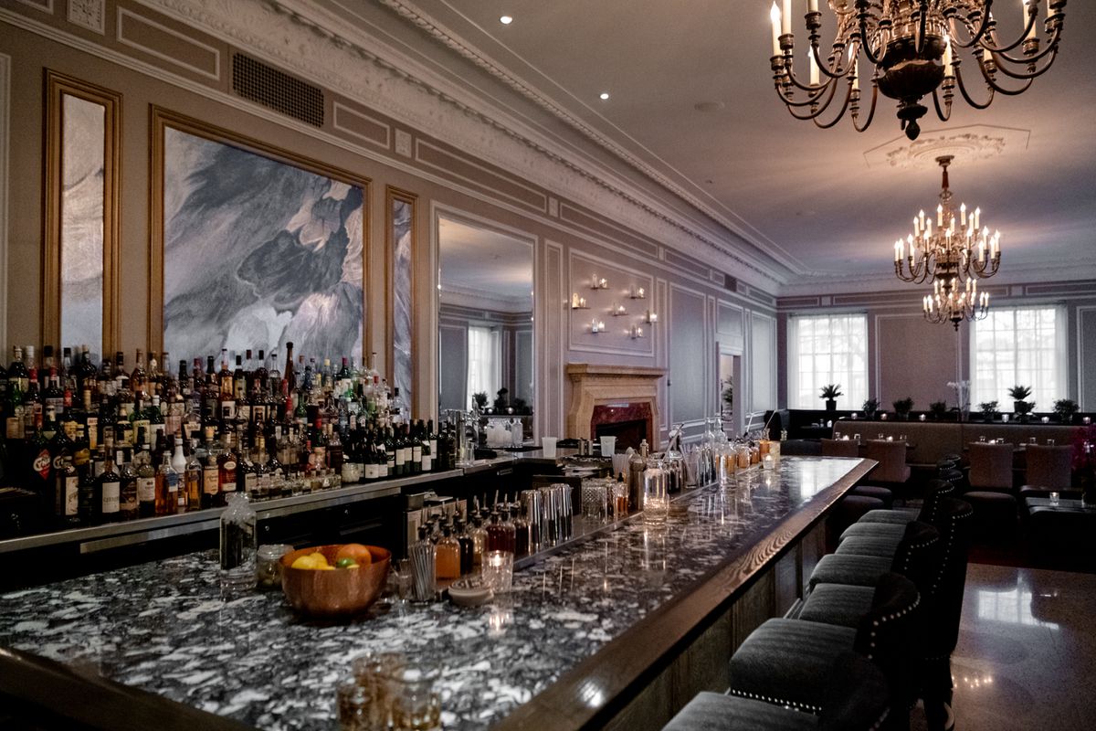 A long gray marble bar with many liquor bars stack behind it. There are mirrors on the wall and chandeliers hanging from the ceiling. 