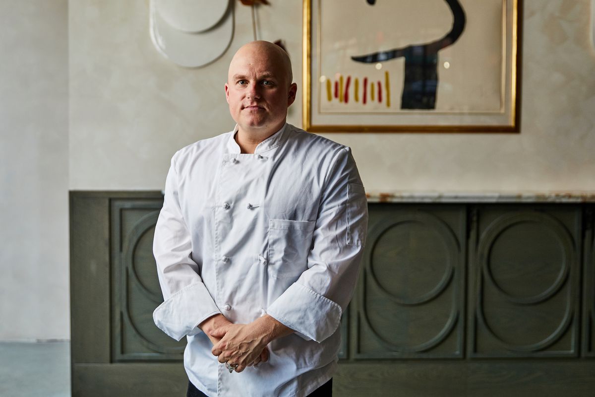 Chef Ryan Bartlow has his hands crossed in front of him, dressed in a white chef’s jacket, standing against a painting in the restaurant