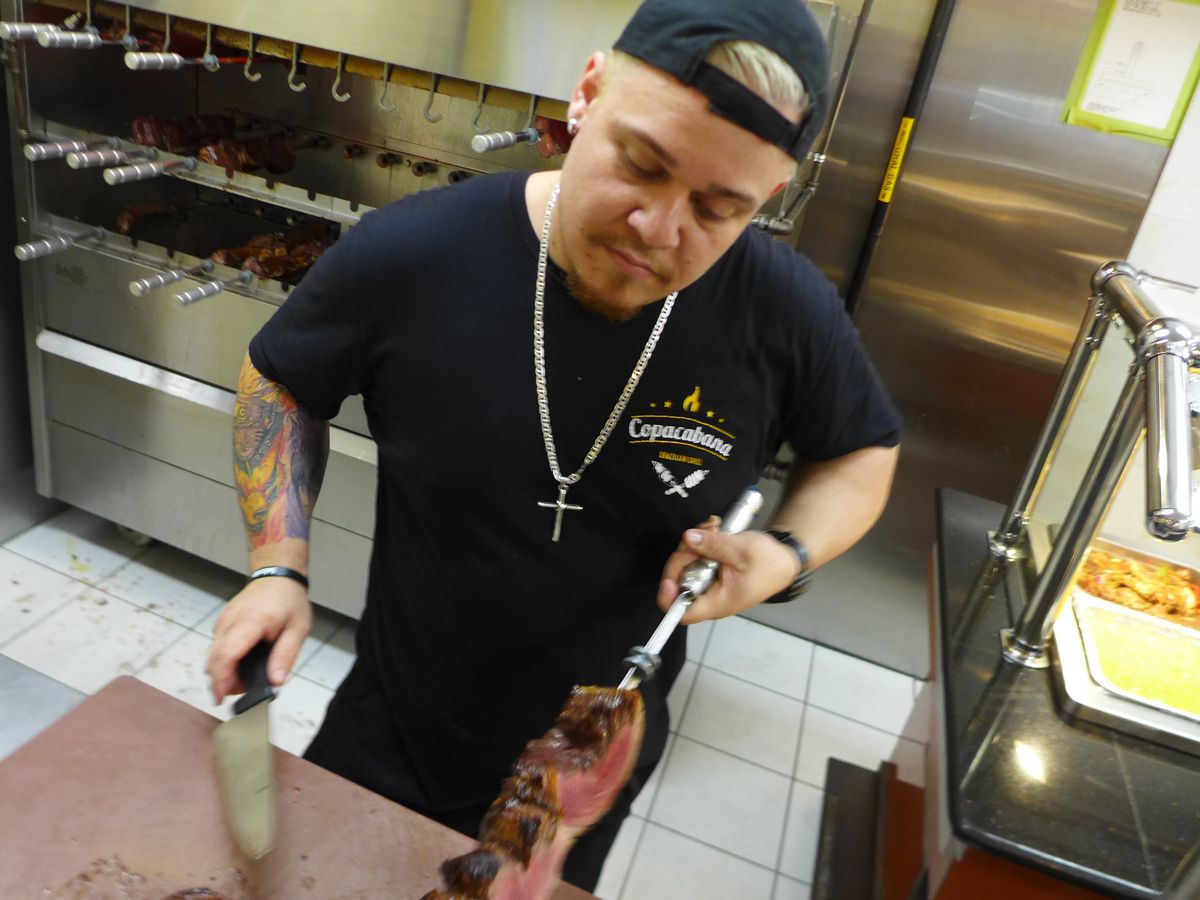 A man in a black outfit with baseball cap turned backwards prepares to carve meat from a long spit pointed downward.