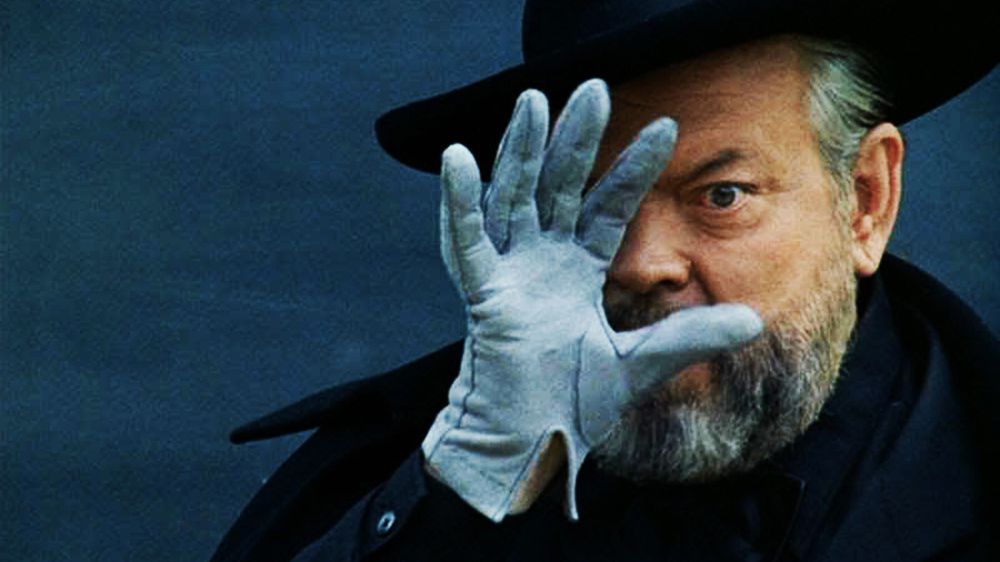 A bearded man (Orson Welles) wearing a black hat, coat, and a white glove holding his right hand against his face.