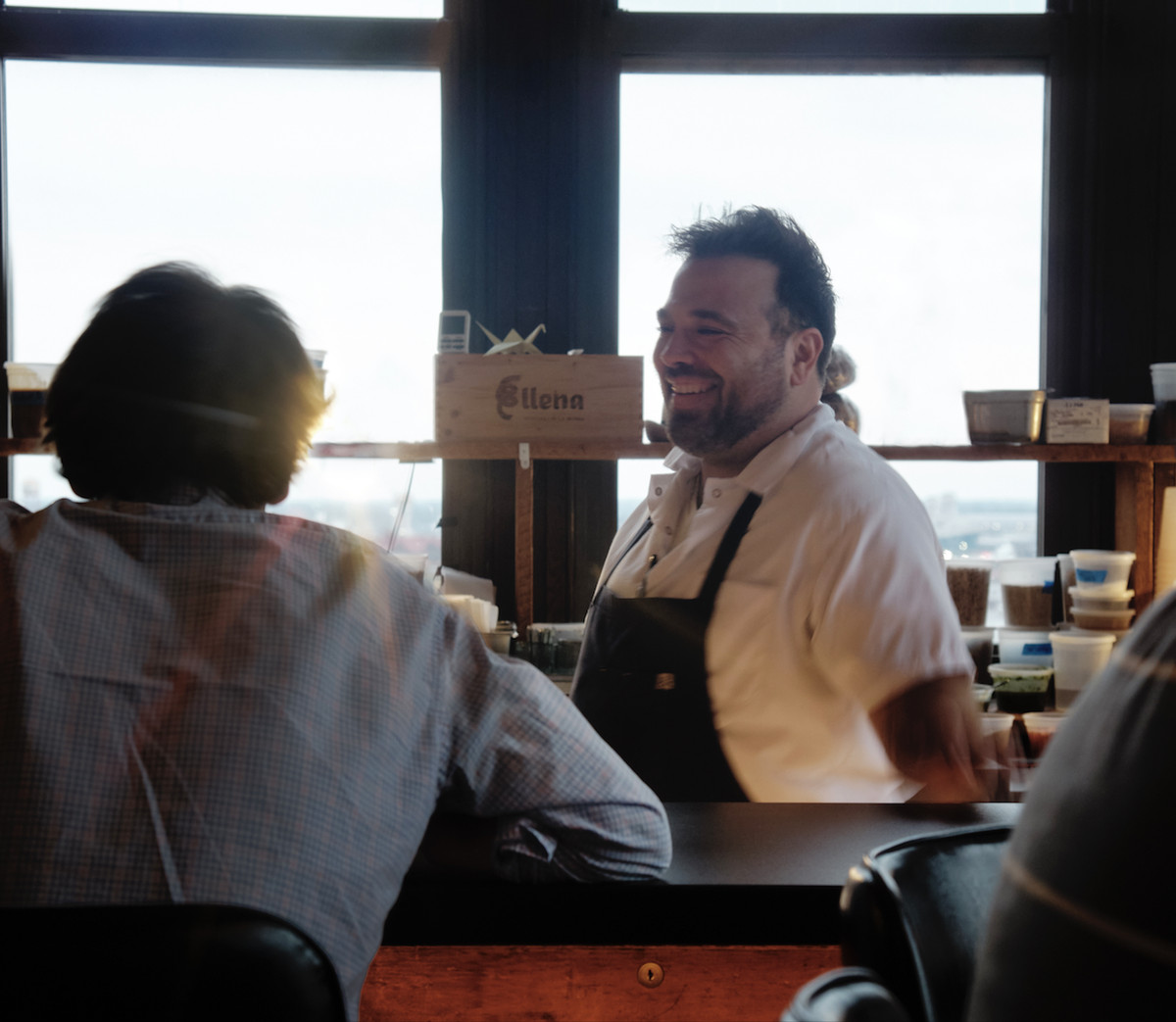 A chef speaking to a diner at a counter in the daytime.