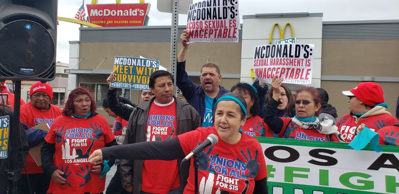 Protesters outside a McDonald’s wear shirts that read “Unions for all, fight for $15” and carry signs that read, “McDonald’s: Sexual harassment is unacceptable” and “McDonald’s: Meet with survivors&gt;”
