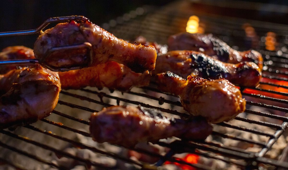 Someone uses tongs to rotate a chicken leg, over a grill with other pieces of basted chicken