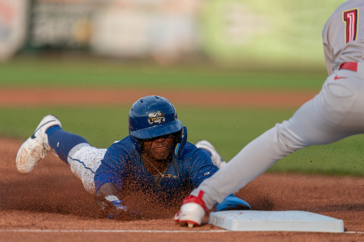 Head-on view of a baseball player in a blue jersey and blue helmet sliding hands-first into third base. Dirt is flying up everywhere and his face is scrunched up from the effort. A defender’s leg juts into the frame, clad in gray pants and white shoes with red accents.