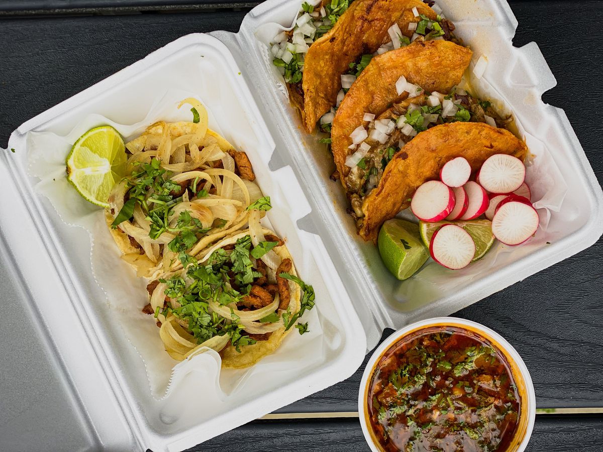 Tacos stacked in a plastic takeout container, with sliced radishes and lime wedges for garnish, beside a paper cup of deep red broth
