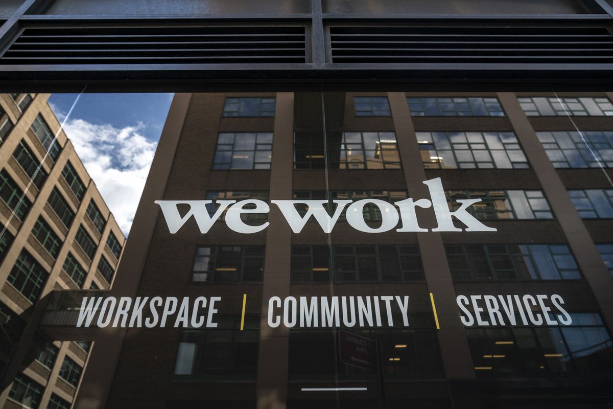 WeWork sign that reads “WeWork workspace, community, services” on a building in DUMBO, Brooklyn, with other buildings and the sky reflected in it.