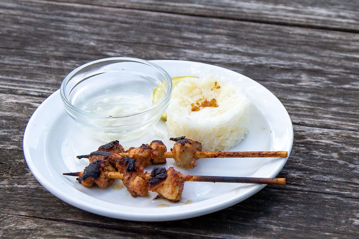 Two skewers of meat, white rice, a clear bowl, a white plate set on top of a wooden surface