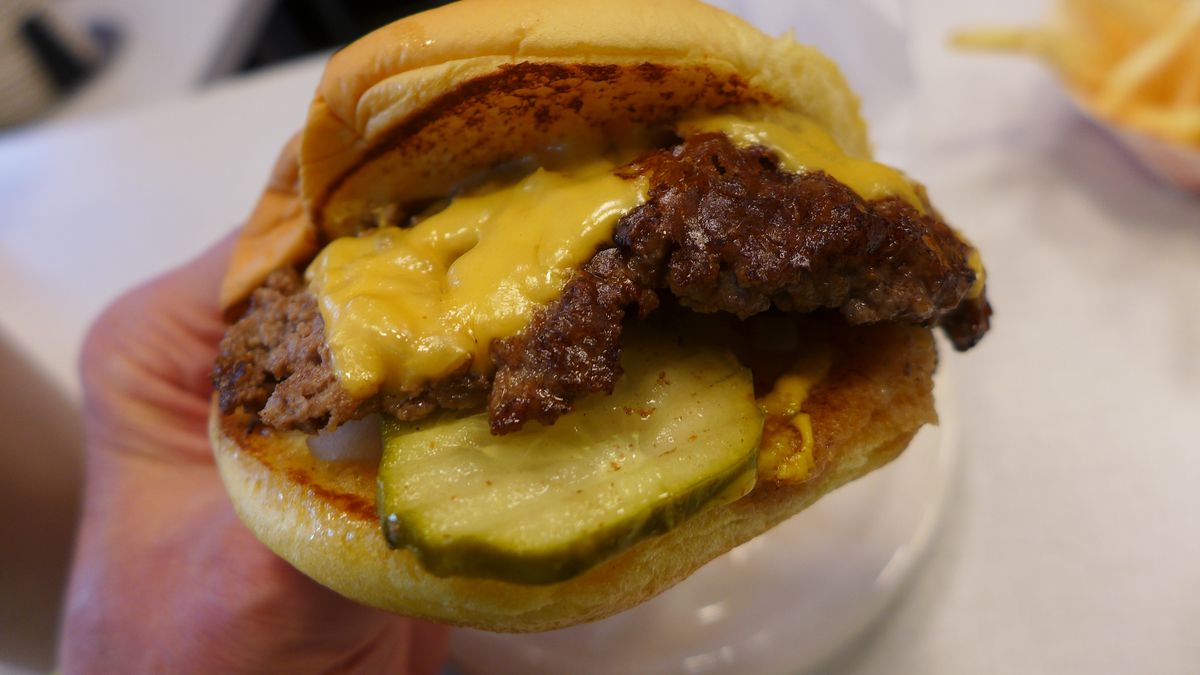 A hand holds a burger aloft, splayed so the ingredients can be seen, patty, pickles, and cheese.