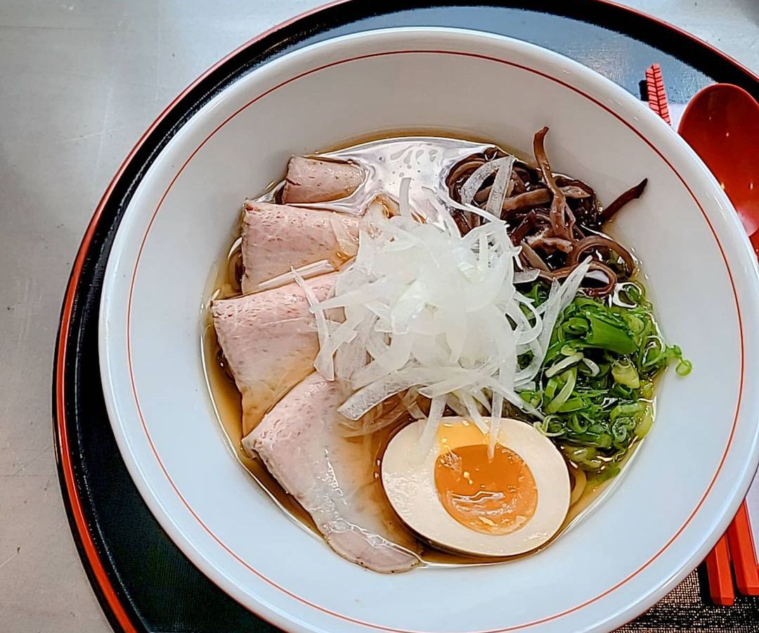 Thin, folded slices of pork sit in a thin brown broth with an egg, greens, and more in a deep white bowl.
