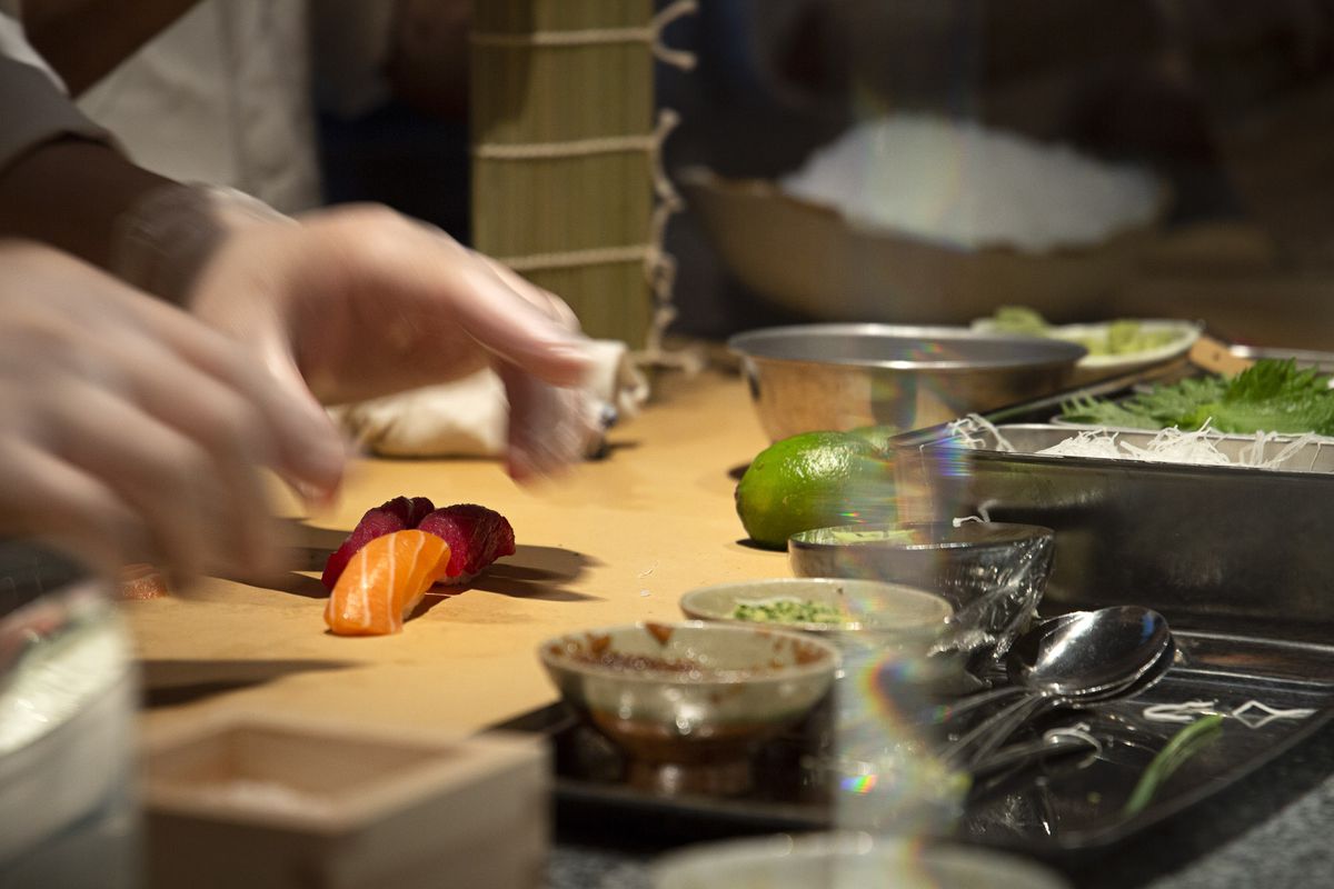Hands reach towards a piece of sushi set on a cutting board surrounded by dishes of indiscernible ingredients
