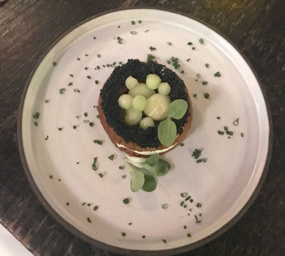 A white plate with a round piece of toast topped with seaweed caviar and small balls of cucumbers with chive scattered around the main dish.