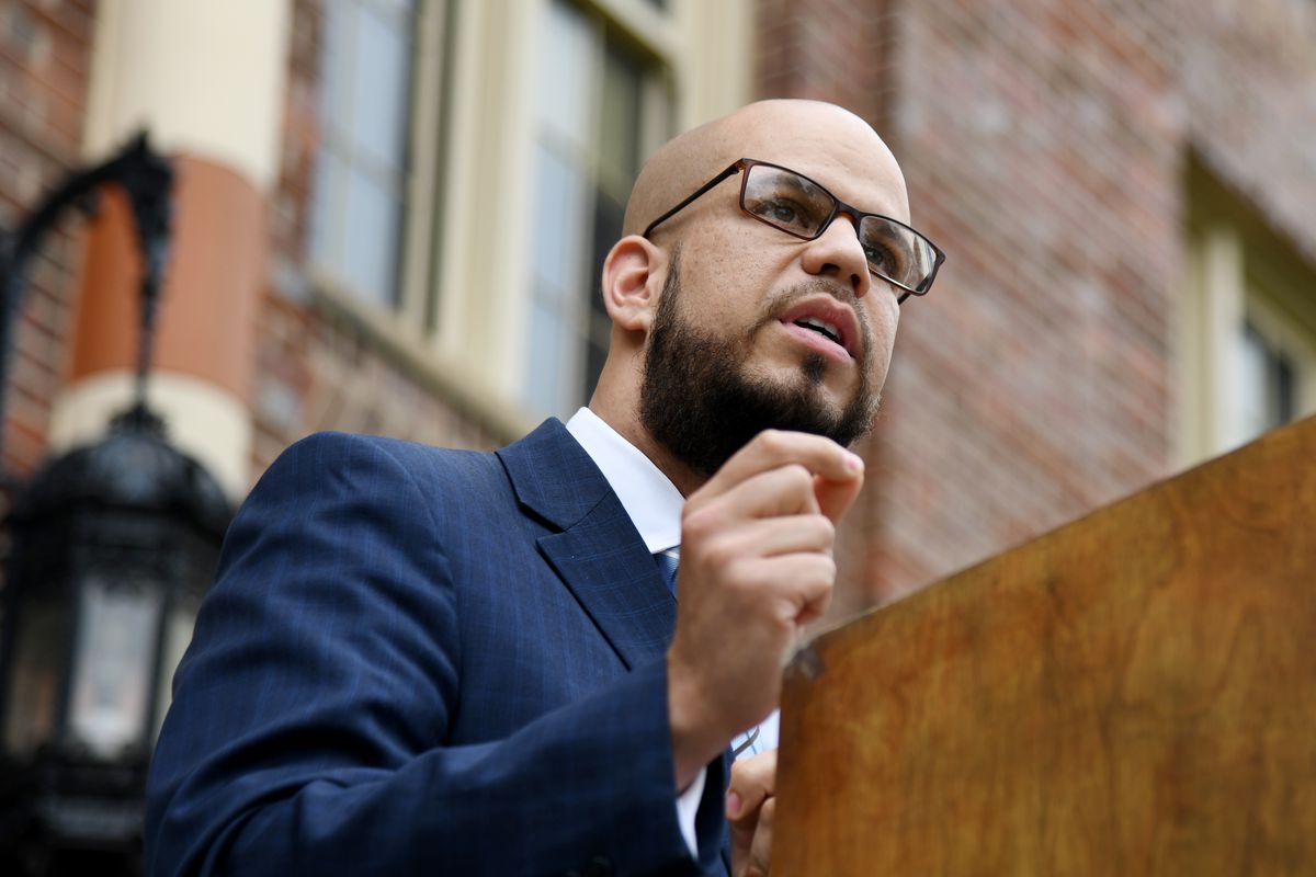Dr. Alex Marrero, wearing glasses and a blue checkered suit jacket, makes a speech at a wooden podium.