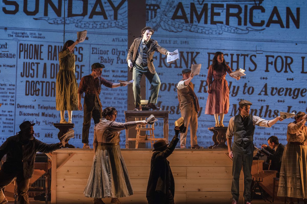 Figures dance on top of a raised wooden platform. One man in the center is spotlit and clutching a newspaper. Images from a historical newspaper are projected onto the background.