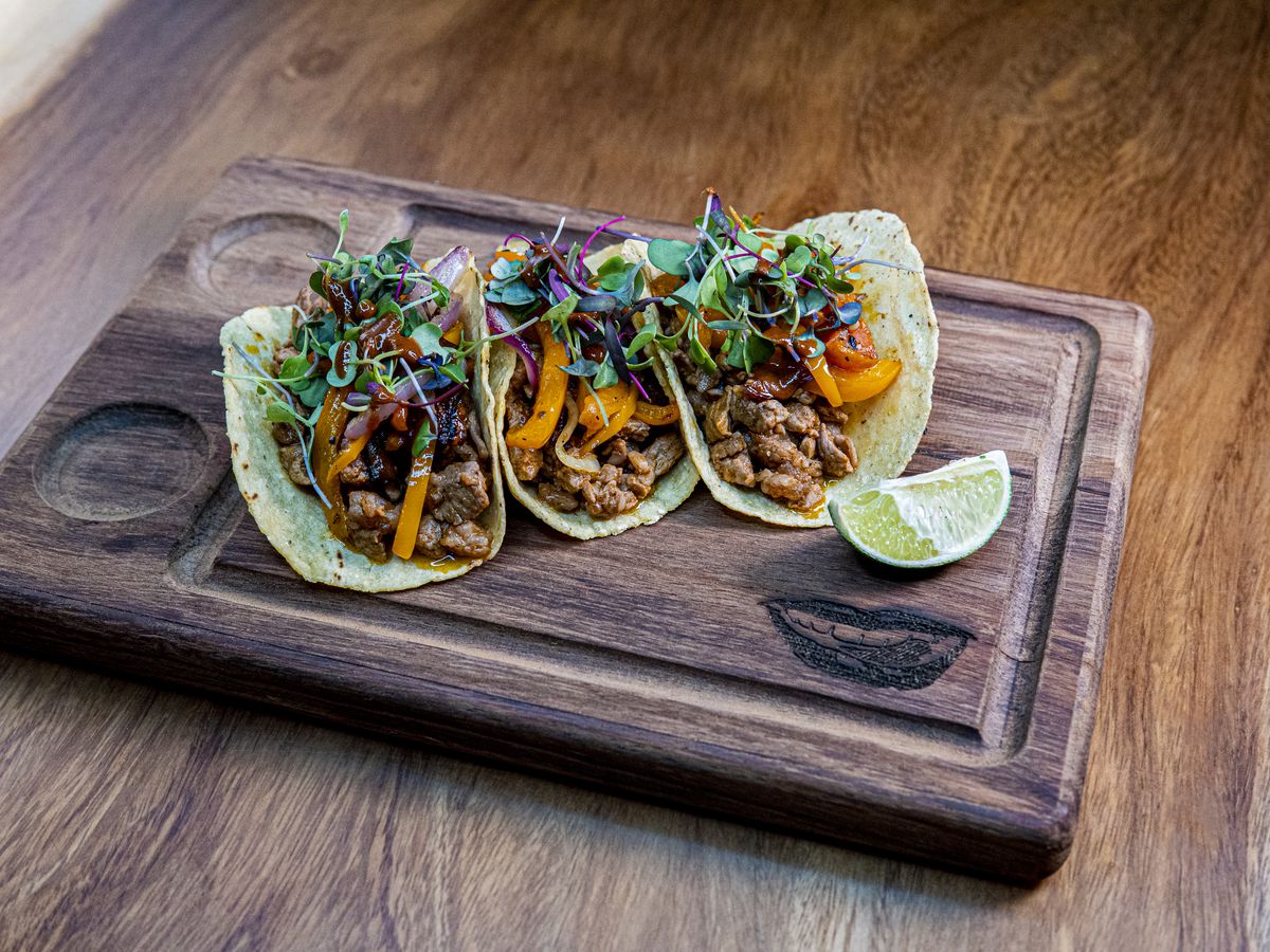 Three neatly plated al pastor tacos atop a wooden carving board from D Boca N Boca in Atlanta.