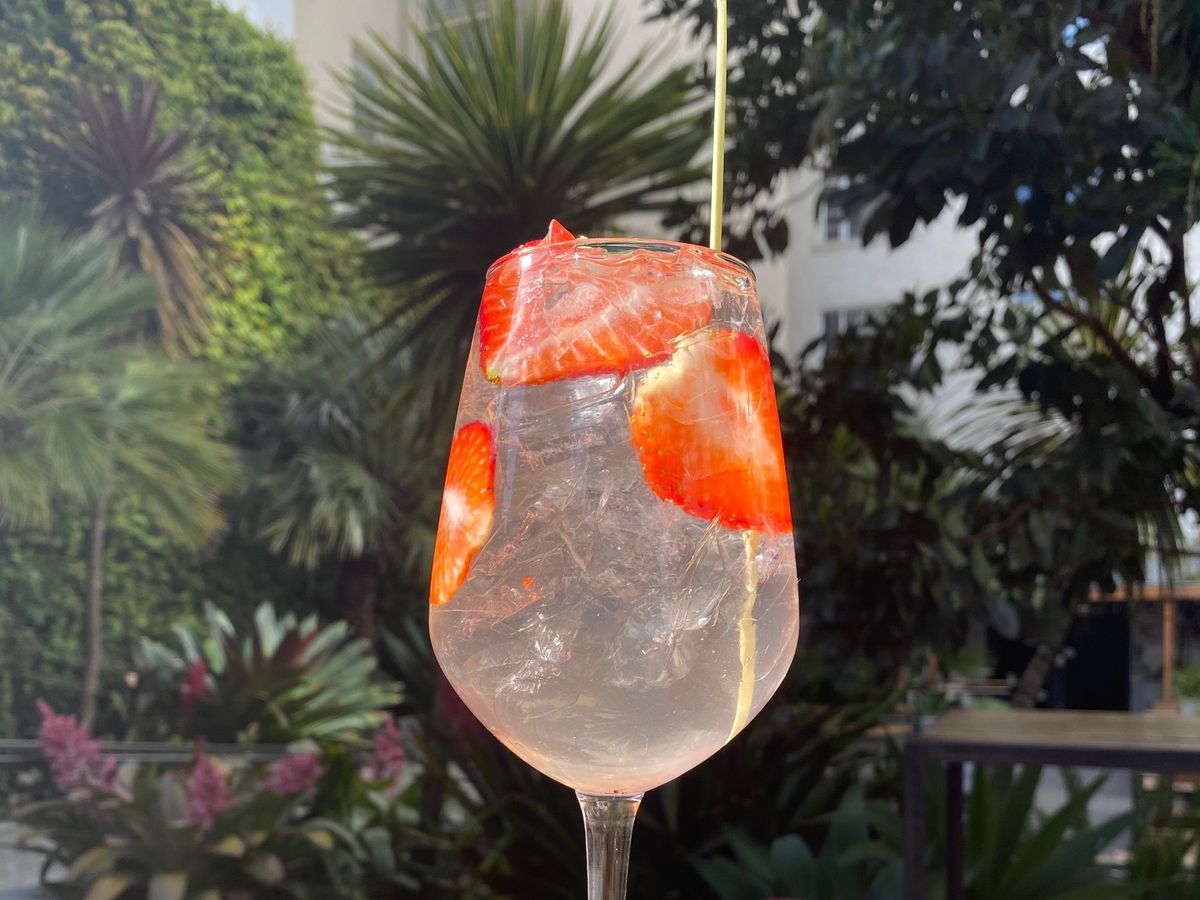 A clear cocktail in a large, stemmed wine glass is sitting on an outdoor table with slices of strawberries floating in the drink.