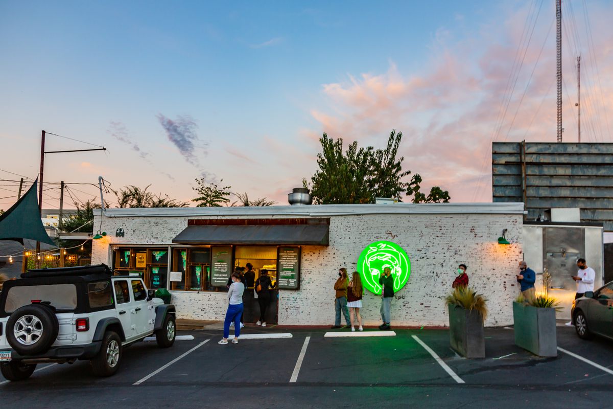 A line of people standing socially distanced and in masks ready to order from Supremo Taco’s to-go window. The shot is taken from the parking lot looking toward the white painted brick building with the bright neon green Supremo logo lit up as the evening sun sets, A white Jeep is parked to the left