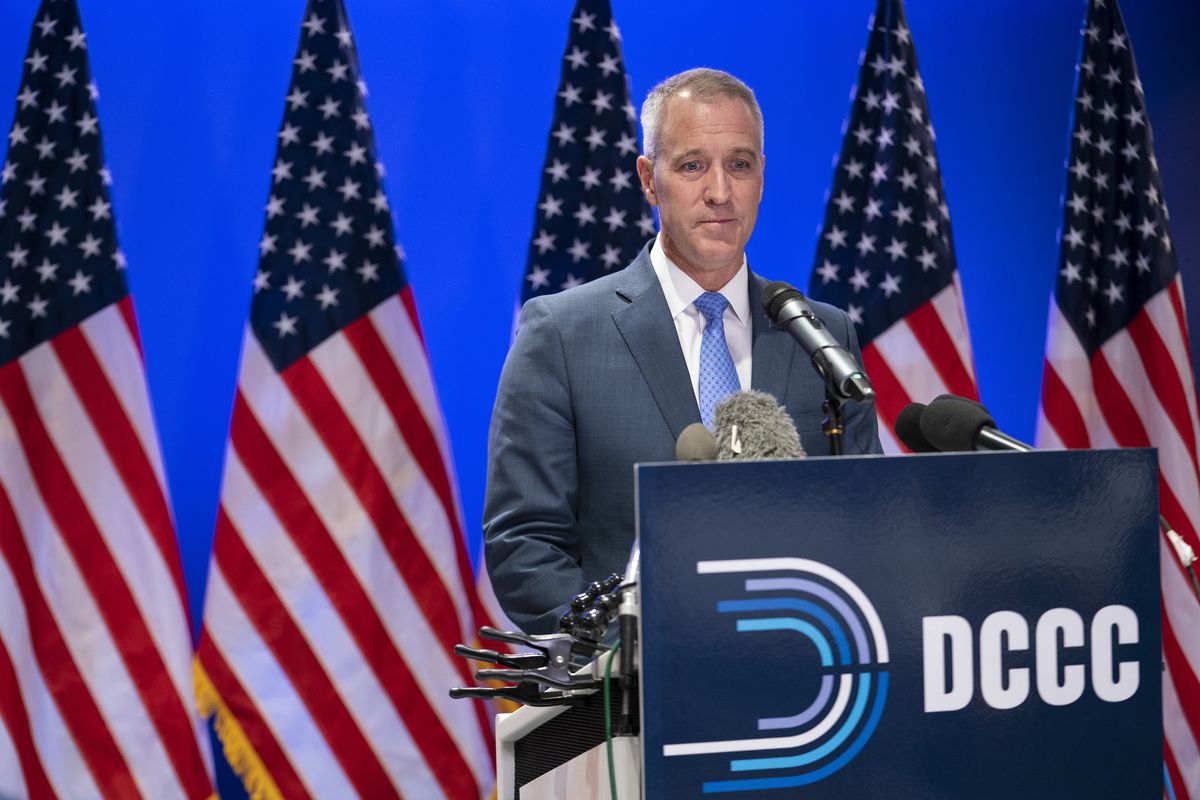 Rep. Sean Patrick Maloney, wearing a blue suit and tie, speaks into a set of microphones at a podium with the DCCC logo on it. Behind him is a row of US flags against a bright blue background.