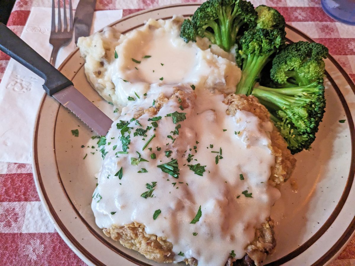 A battered and fried piece of meat with gravy, mashed potatoes, and steamed broccoli.