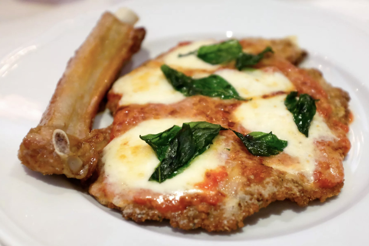 a white plate of veal parmesan covered in red tomato sauce, gooey white cheese and basil leaves. next to the veal is a bone of some sort.