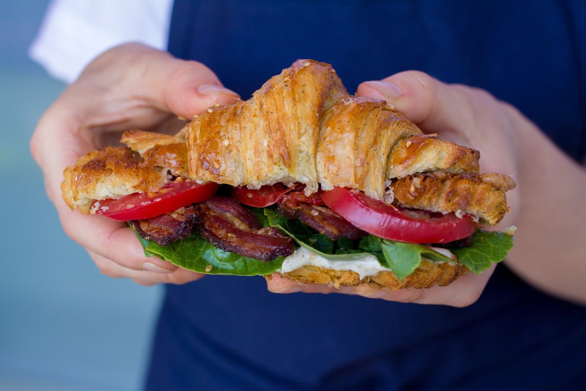 A view of hands holding a BLT on a croissant, with lamb bacon.