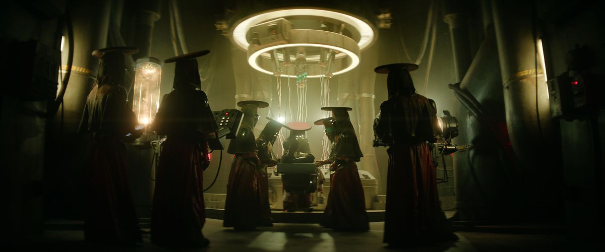 A group of mysterious figures in red robes, flat-topped hats, and veils surround an object under a circular, ringed device hanging from the ceiling in Rebel Moon Part One:&nbsp;A Child of Fire
