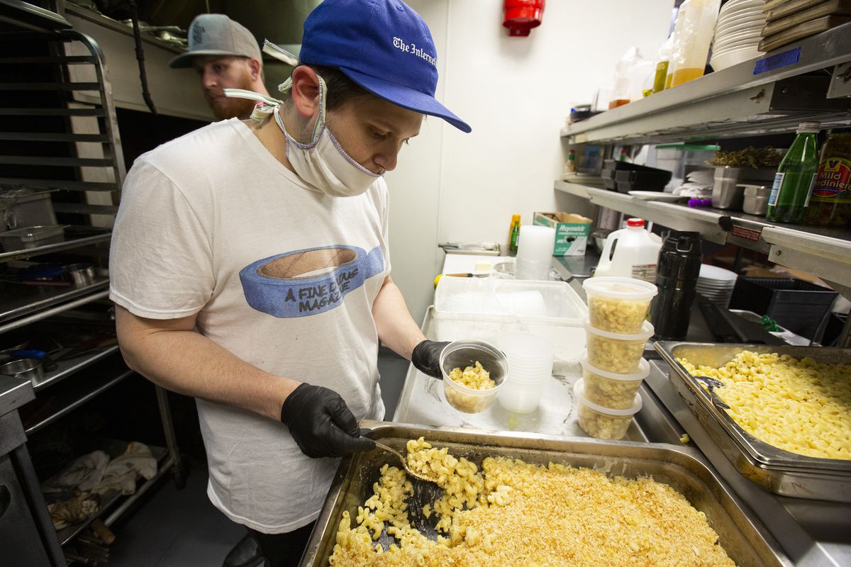 A man wearing a medical mask scoops mac and cheese into plastic containers.