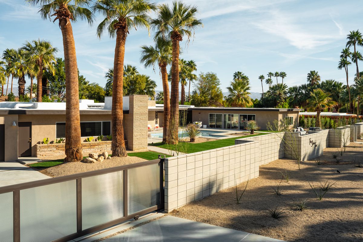 An external view of a ranch midcentury modern house, with an automatic gate, brick wall, and palm trees around. 
