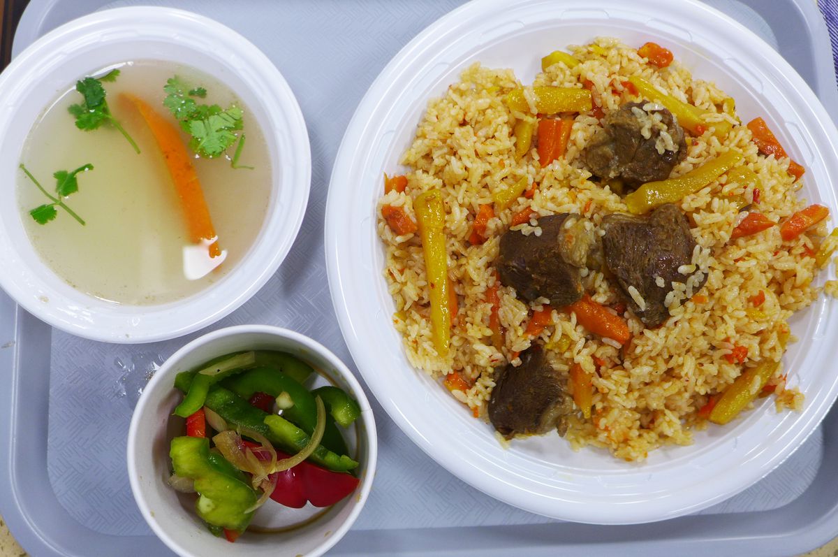 A plate of orange rice dotted with lamb, a soup floating a carrot, and a bell pepper salad, all on a cafeteria tray.