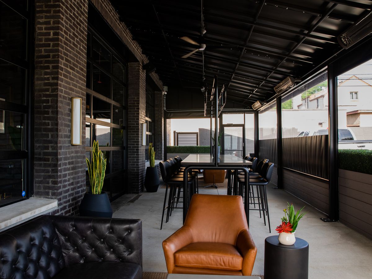 A dark-colored leather sofa to the left, a lighter-colored upholstered seat, a small round end table with a potted plant, in the background more seating and table space in an enclosed patio at Petty Cash in Detroit, Michigan. 