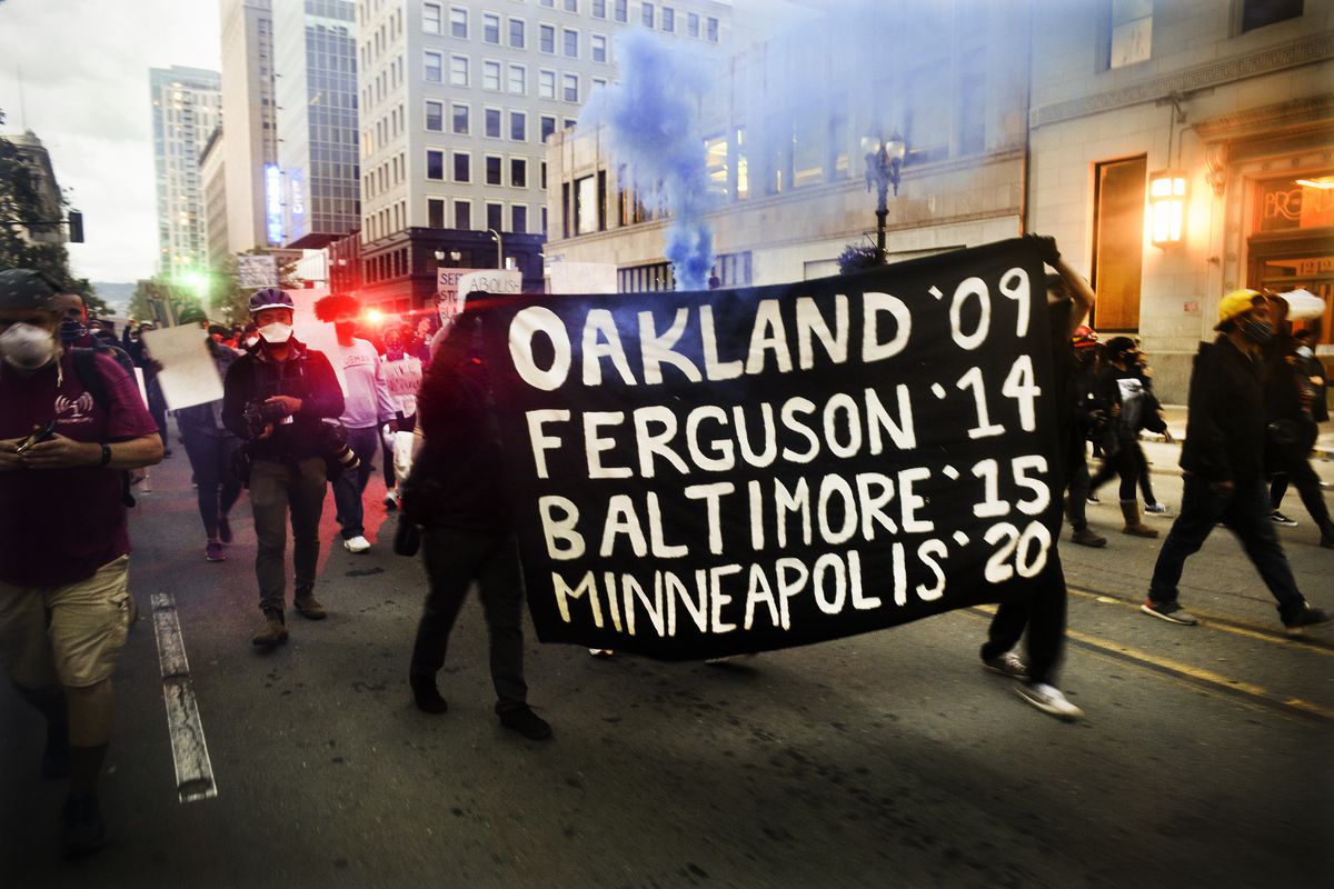 Amid shining traffic lights, smoke, and a sinking sun, a group of protesters march through the street. They are led by two people in black, holding a black flag with bold white text that reads, “Oakland ’09 Ferguson ’14 Baltimore ’15 Minneapolis ’20.”