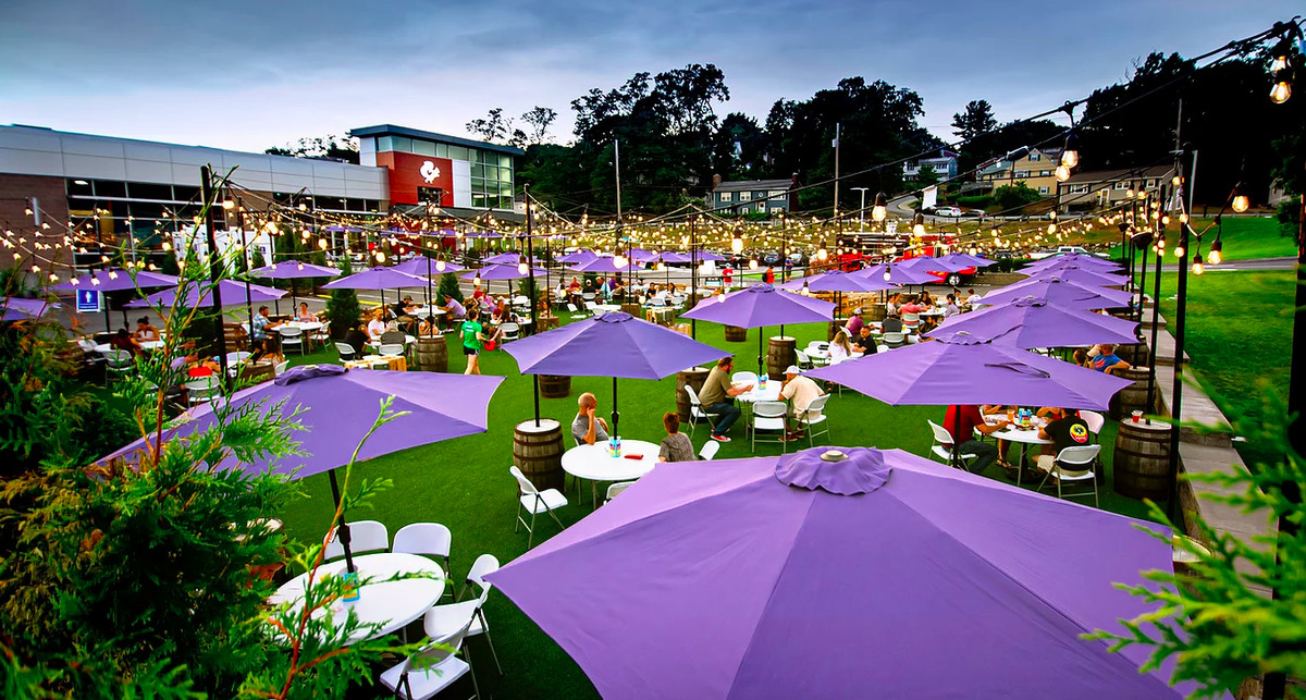 An expansive brewery patio features green turf and lavender umbrellas over round white tables