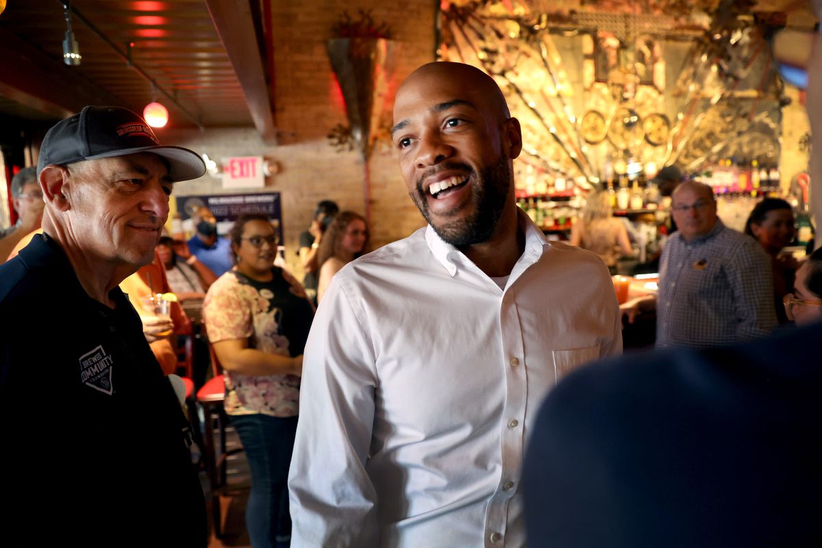 Barnes, a Black man in a white Oxford shirt, smiles broadly amid a mostly white crowd of supporters. Behind him is a golden-lit bar.