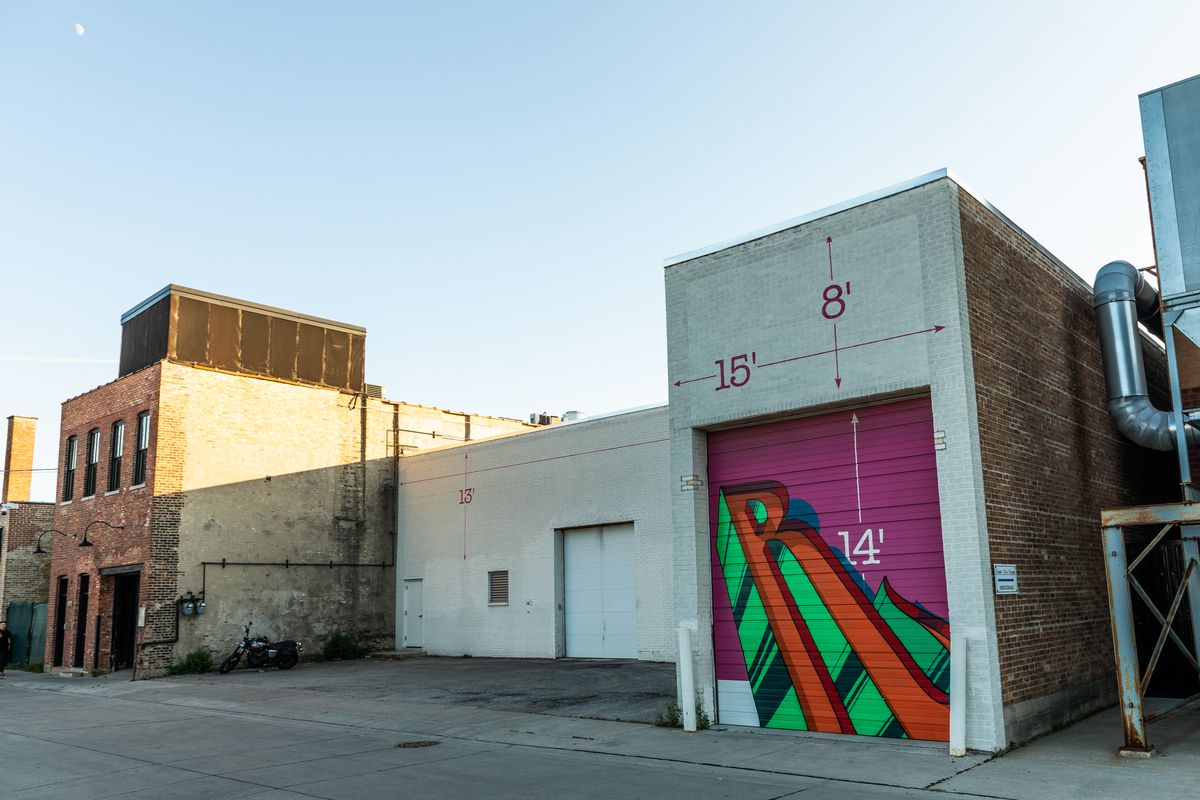 A row of old warehouse buildings stand jagged together. The closest brick building with a metal rolldown garage door has a red and green graffiti “R” against a pink background.
