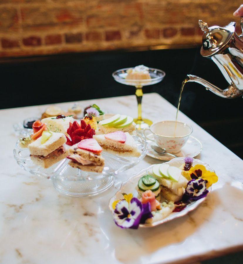 A teapot pouring tea into a decorative mug, alongside a platter of colorful sandwiches and sweets, on a marble dining table surrounded by a banquette 