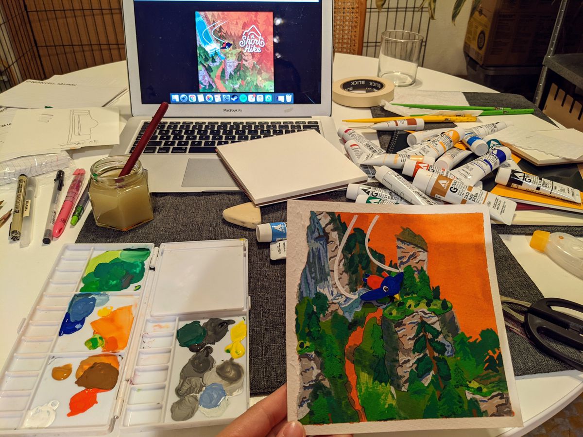 A messy table with lots of paint bottles strewn across. In the background a laptop displays an art image for the game A Short Hike. In the foreground, a paint palette has lots of colors on it, and a painting of a Short Hike is next to it.