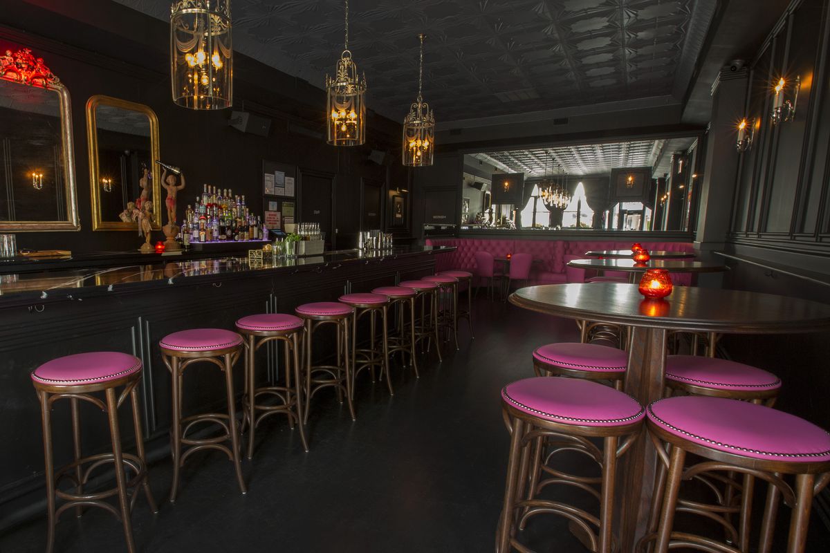 A bar with black walls, vintage gold mirrors and light fixtures, plus pink stools.