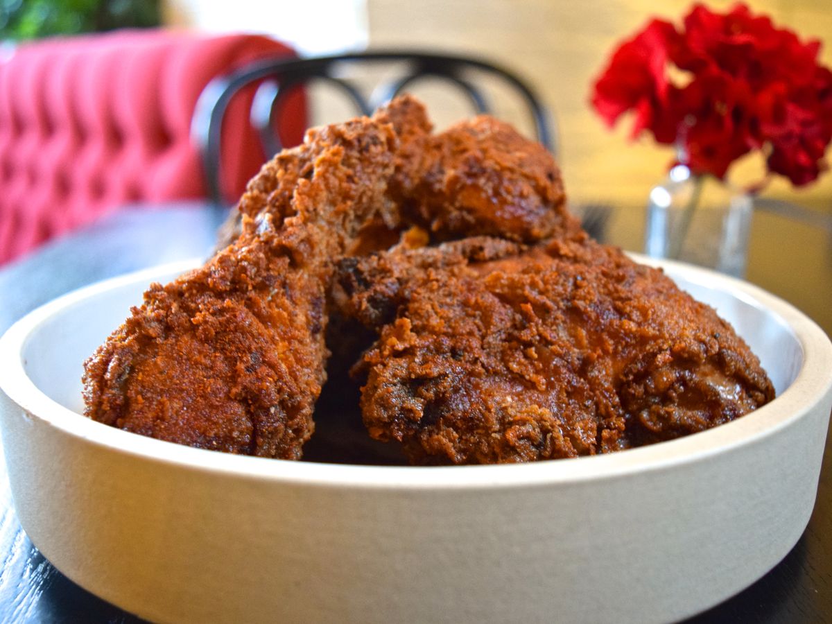 Several pieces of fried chicken sit in a high-rimmed white bowl on a table with a vase of red flowers visible in the background. Plush red booth seating is also visible.