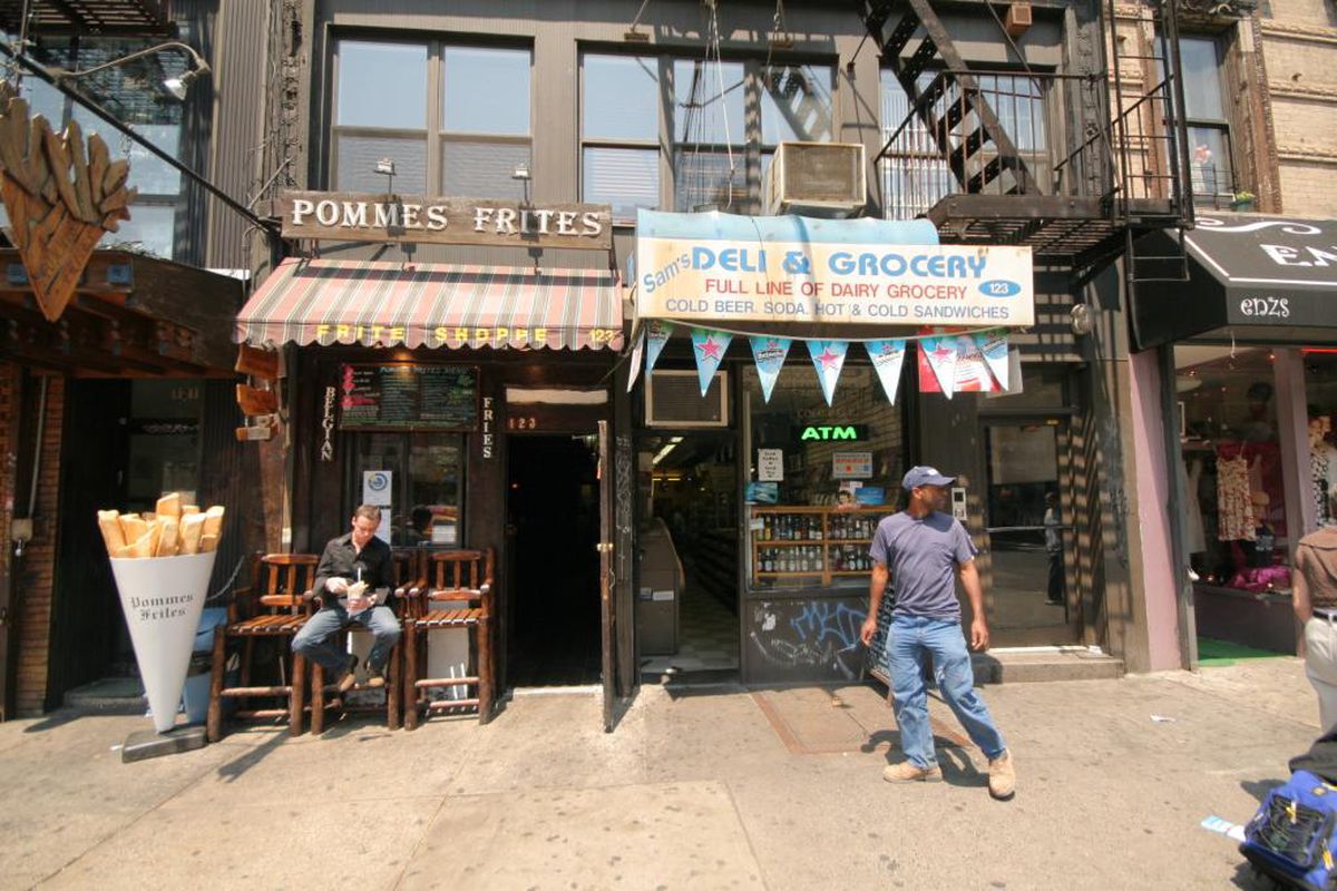 The exterior of restaurant Pommes Frites. A man is sitting on a bench in front of it eating, another man is walking by.