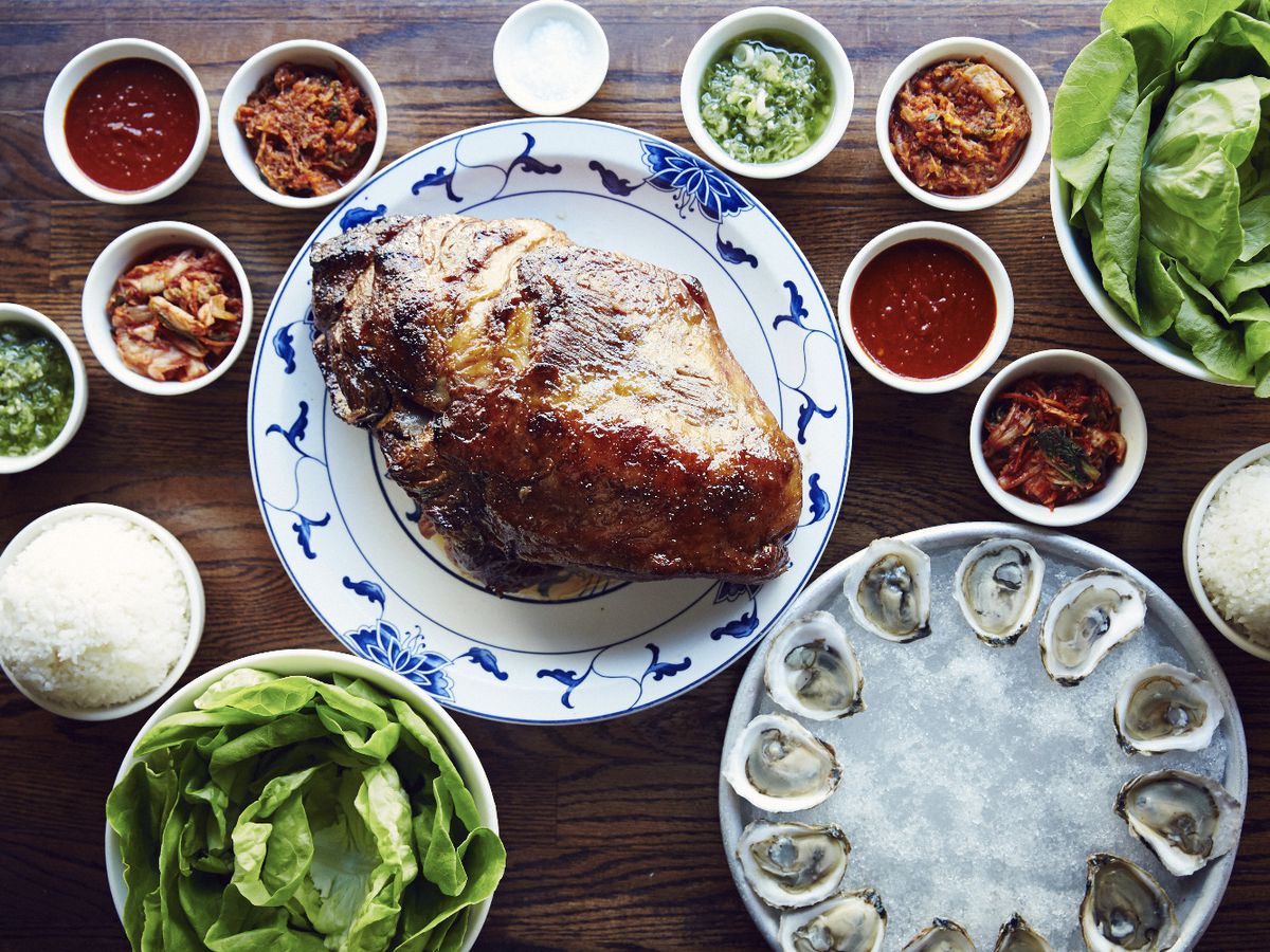 The bo ssäm pork butt at Ssäm Bar sits surrounded by oysters on the half shell, multi-colored sauces, and lettuce leaves