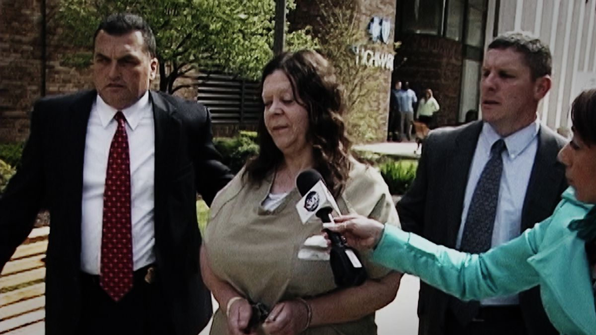 A woman in a tan shirt and handcuffs flanked by two large men in suits being interviewed by a woman in a turquoise blazer holding a microphone.