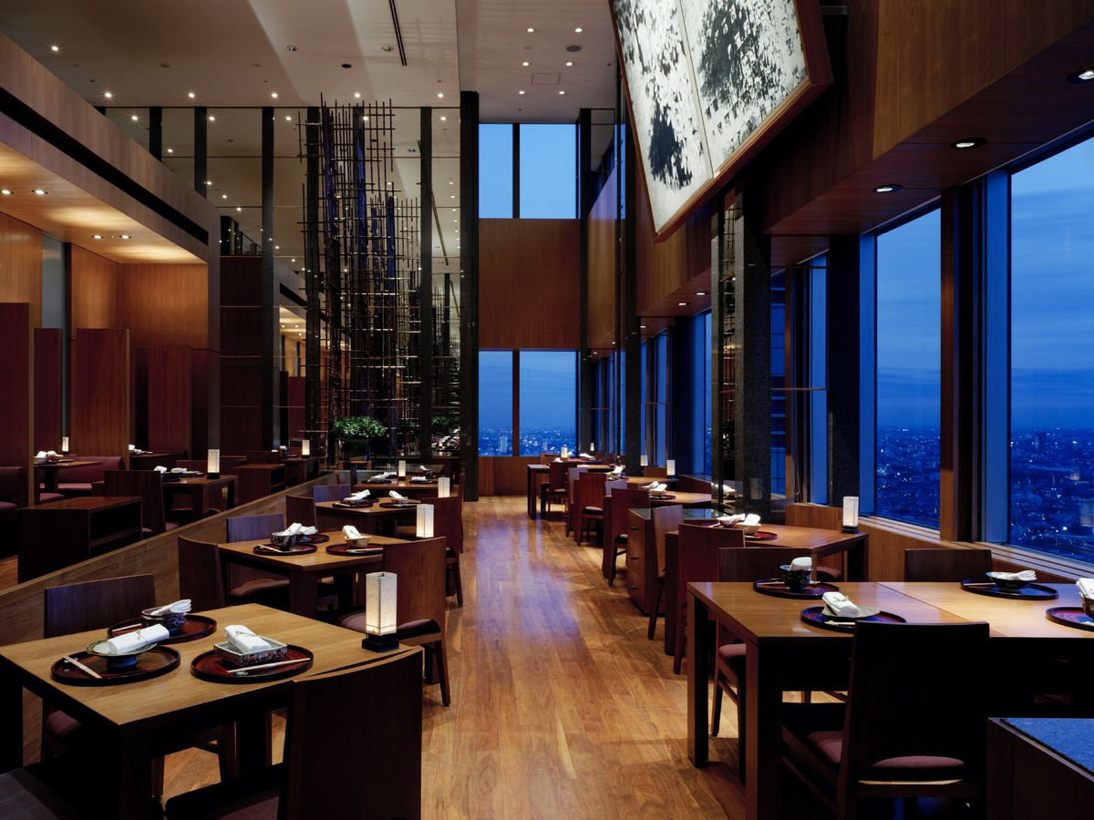 A restaurant interior with high ceilings, wood floors, a city view out the window in the evening, and tables set for dinner. 