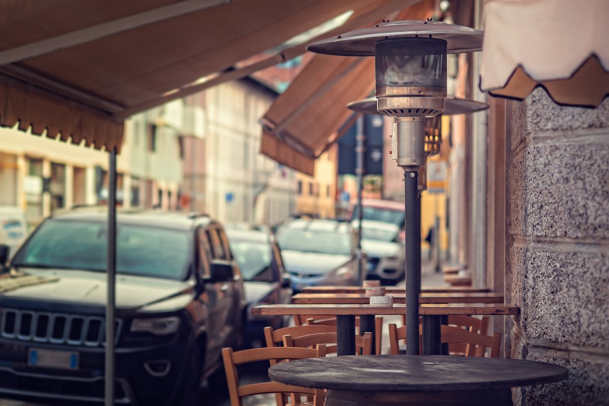 An unlit propane heater standing outside on a street sidewalk with restaurant tables and chairs in the background.