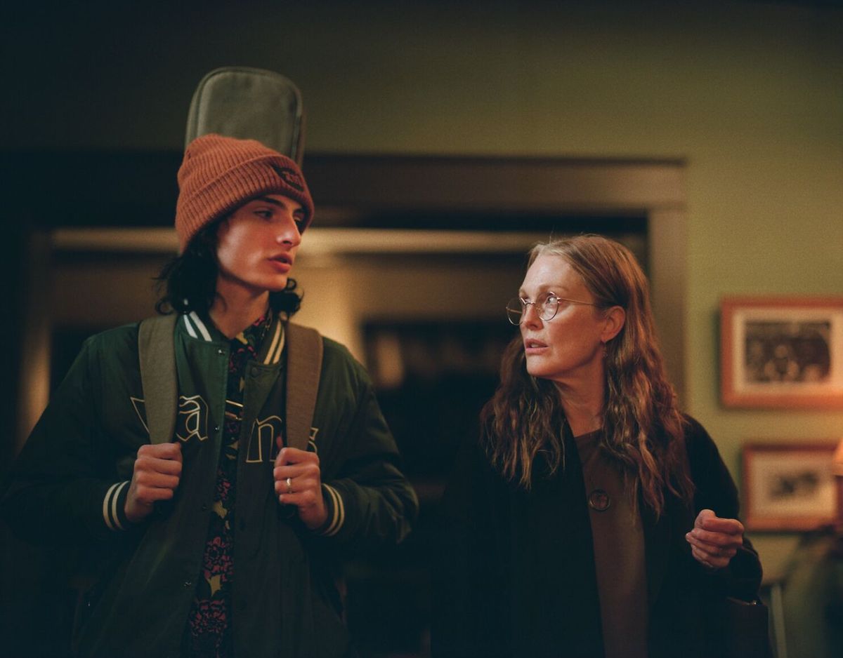 A teenage boy (Finn Wolfhard) with a guitar case on his back stands next to an older, long-haired woman (Julianne Moore) with glasses.