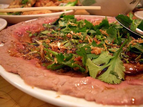 A close-up on a dish nearly covered in a thin layer of beef in a light sauce topped with sprigs of greens and crunchy garnishes.