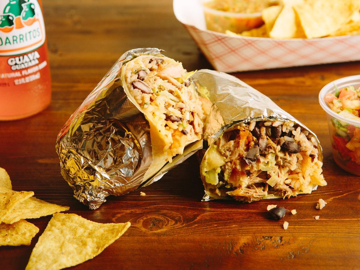 A burrito, split in half and wrapped in foil, sits on a wooden table with a side of salsa and tortilla chips nearby. An orange Jarritos soda bottle is visible in the background.