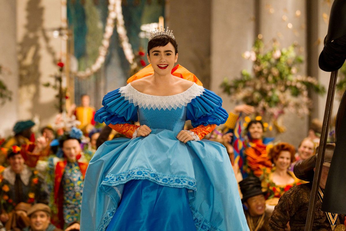 Snow White (Lily Collins) ascends the throne room stairs in a lavish blue gown with white trim and a giant orange bow on her back as people dressed in equally colorful costumes cheer her own