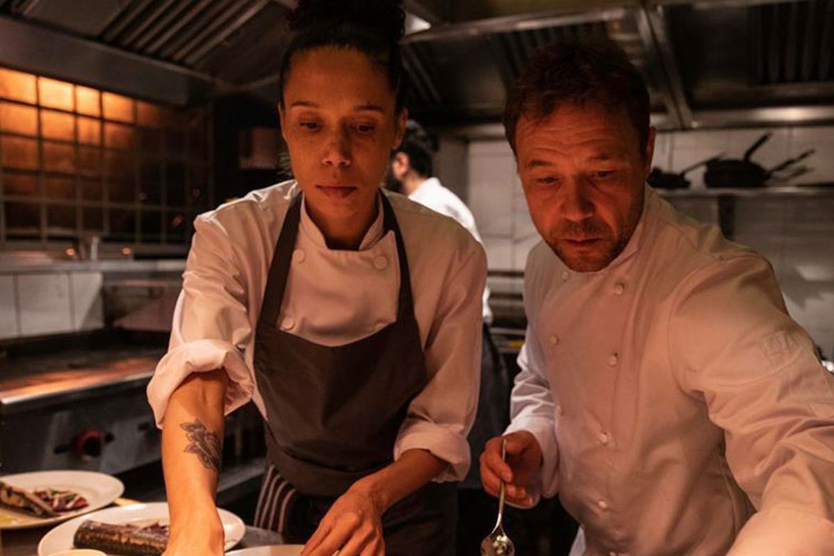 Two chefs in whites stand over the pass in restaurant film Boiling Point. On the left is Carly, played by Vinette Robinson, and on the right is Andy, played by Stephen Graham.