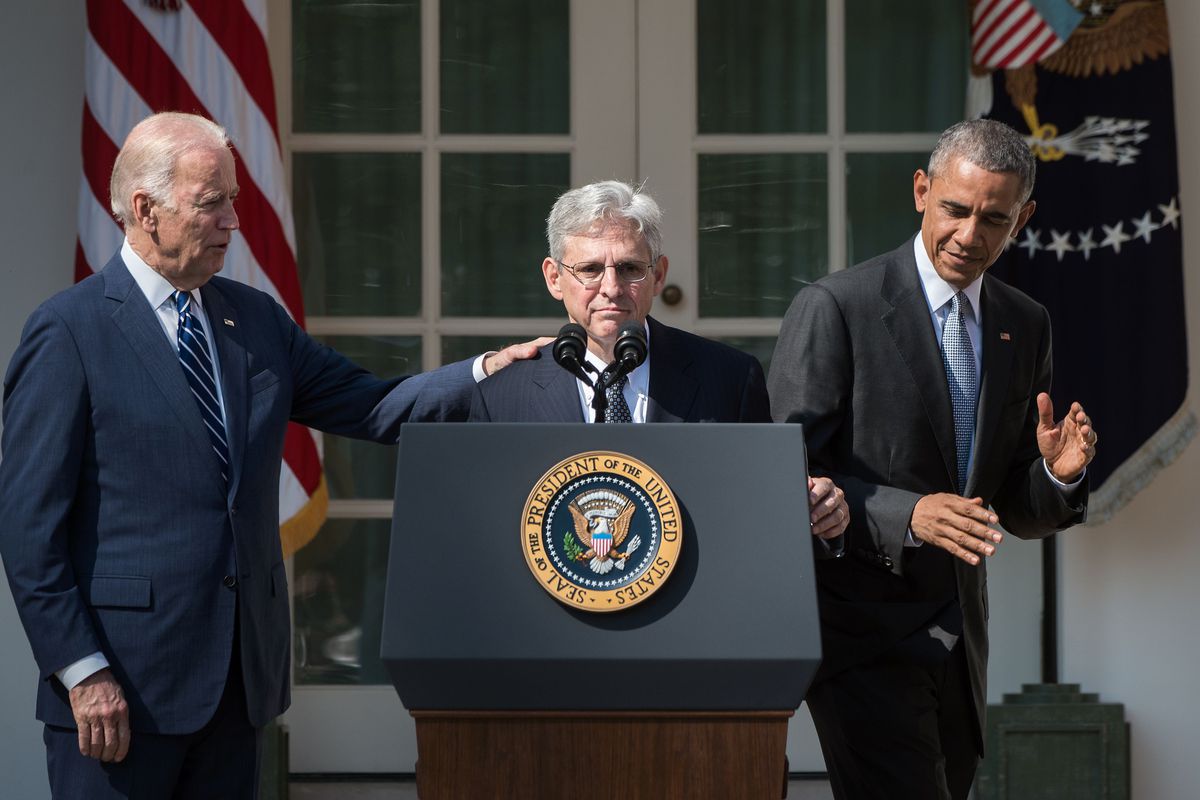 Merrick Garland stands at a lectern. Vice President Joe Biden stands with his hand on Garland’s shoulder. President Barack Obama stands on Garland’s other side.